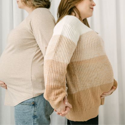 Two pregnant women stand back to back, holding hands