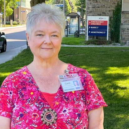 A mid shot image of Carol Foley who has short grey hair and blue eyes, and is wearing a bright pink top.