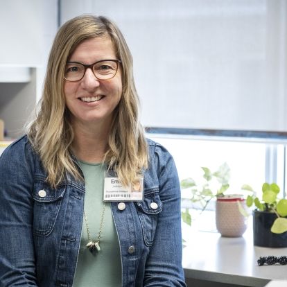 Emily Jackson has shoulder-length blonde hair and blue eyes, and wears glasses. She’s wearing a green top with a jean jacket and is pictured sitting at her desk at KidsInclusive at Hotel Dieu Hospital.