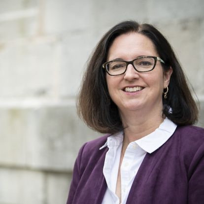 Dr. Jocelyn Garland has shoulder length, dark hair. She's wearing a purple jacket with a white collared shirt. She also wears glasses. She photographed outside the Kingston General Hospital site.