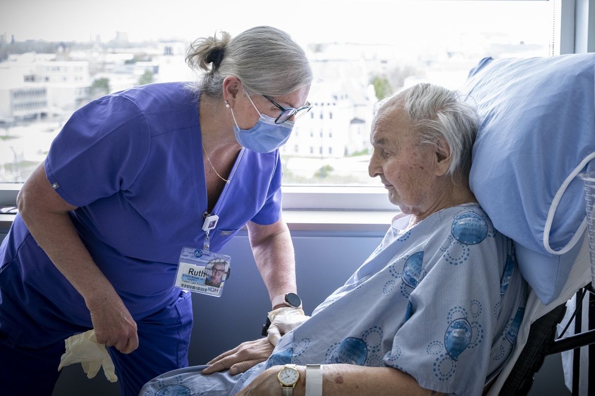 Ruth Bradshaw is pictured caring for a patient in a wheelchair in their room. Bradshaw has white/silver hair, pulled back into a bun, black framed glasses and is wearing blue scrubs with a mask. She’s leaning over the patient and her hand is on his arm in a loving way.