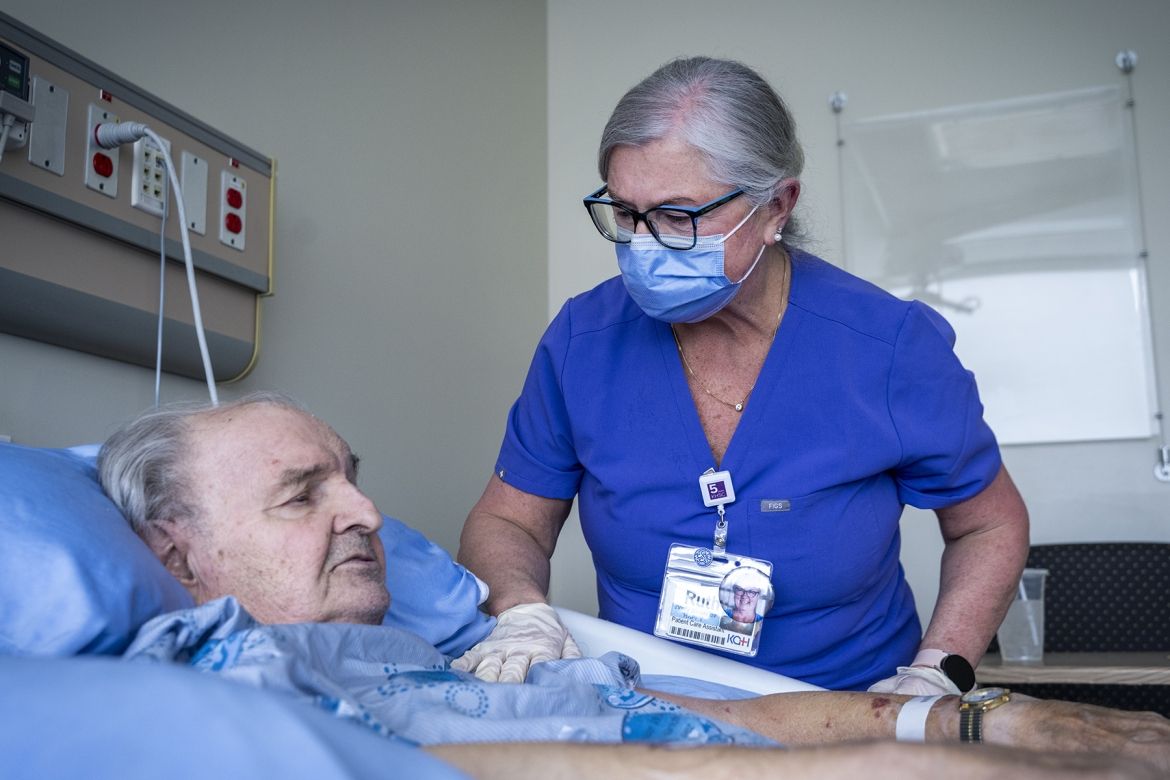 PoKHSC - Ruth Bradshaw Ruth Bradshaw is pictured caring for a patient who is lying in a hospital bed. Bradshaw has white/silver hair, pulled back into a bun, black framed glasses and is wearing blue scrubs with a mask. She’s leaning over the patient in the bed, and her hand is on his upper arm in a loving way.