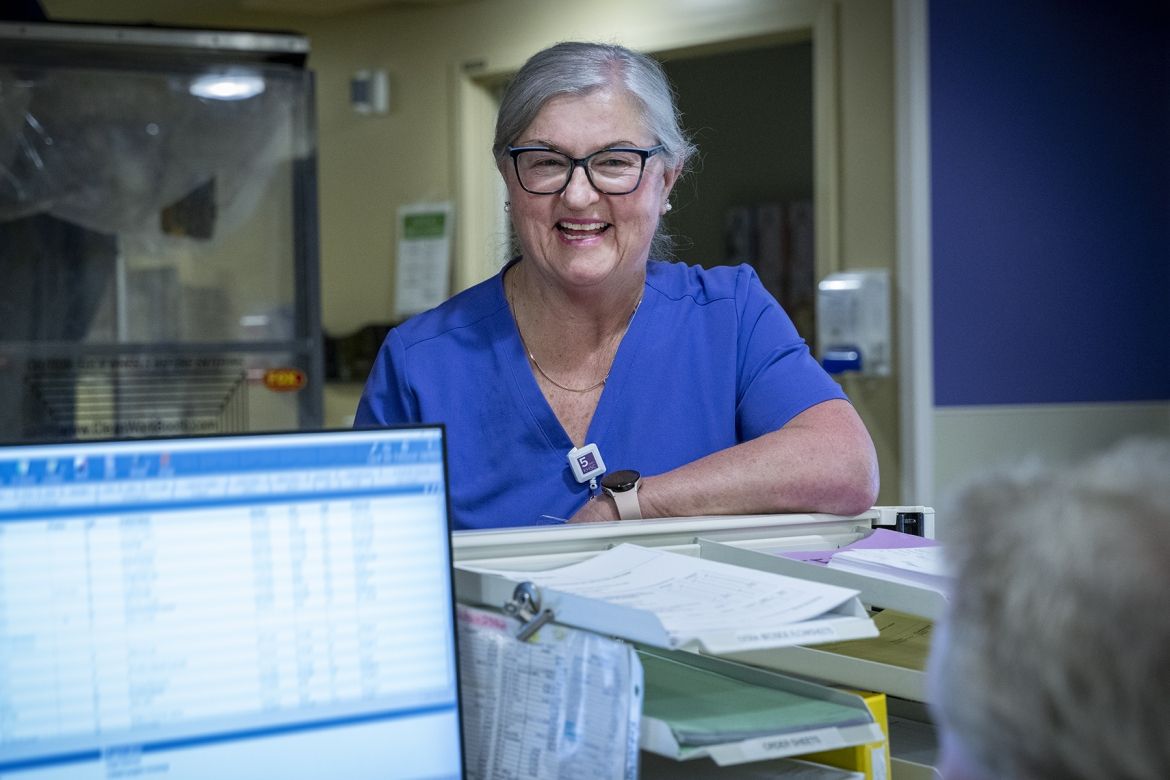 Ruth Bradshaw is pictured smiling and chatting with a colleague, while standing behind a care desk at Kingston General Hospital. She has white/silver hair, pulled back into a bun, black framed glasses and is wearing blue scrubs.