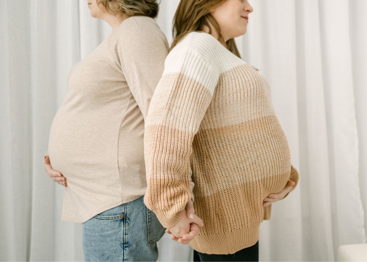 Two pregnant women stand back to back, holding hands