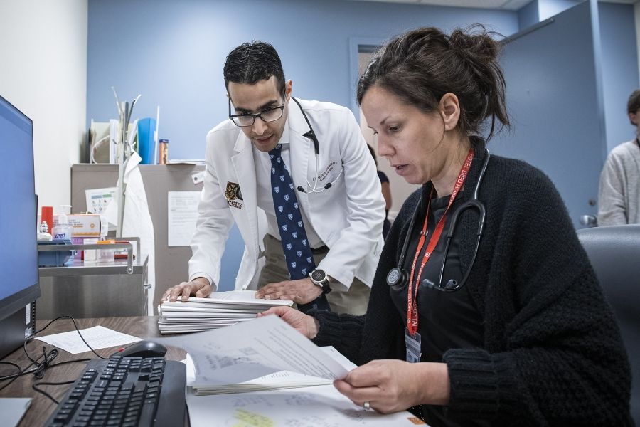 Dr. Almufleh is pictured hovering and chatting with the colleague, who is sitting at a desk in front of a computer. The duo is looking over paperwork.