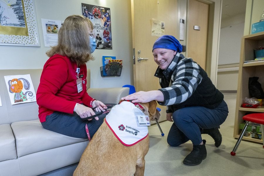 People of KHSC - Julie Halle + Doc Julie Halle and Dog interact with a KHSC patient. The patient is crouching as she pets doc smiling. Julie is facing the patient and chatting.