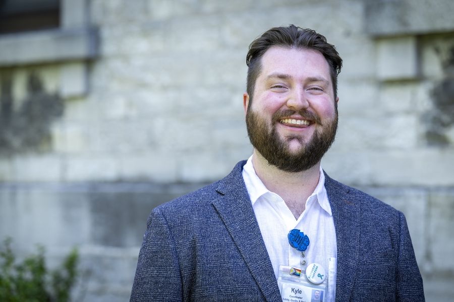 A man is standing outside Kingston General Hospital smiling directly at the camera. He has brown hair, slicked back and brown eyes. He’s wearing a blue plaid blazer over a white collared shirt.