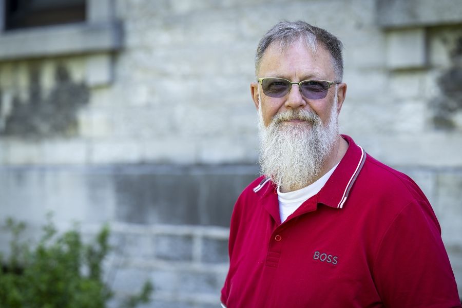 A man is standing outside Kingston General Hospital smiling directly at the camera. He has short, salt and pepper hair, is wearing transitions glasses and had a long white beard. He’s wearing a bright red collared top over a white t-shirt.