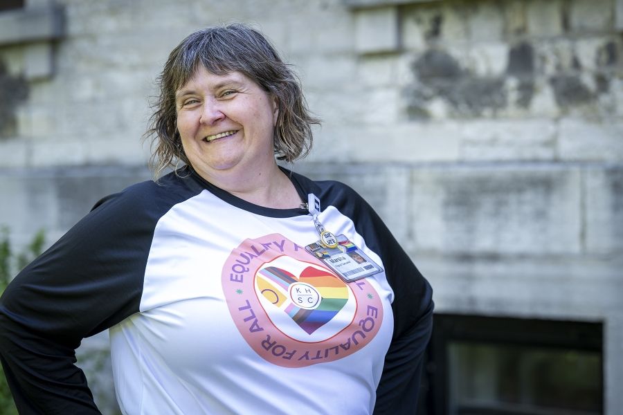 A woman is standing outside Kingston General Hospital smiling directly at the camera. She has short, brown wavy hair, and brown eyes. She’s wearing a KHSC pride long-sleeve t-shirt that says equality for all.