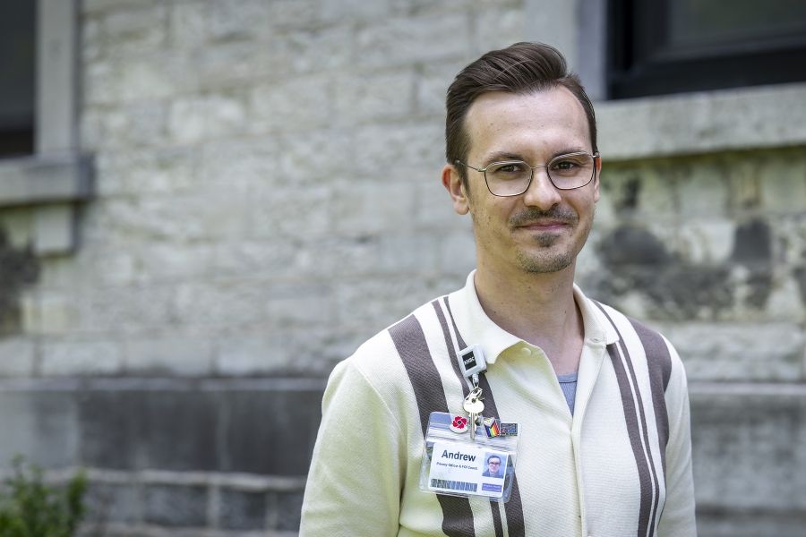 A man is standing outside Kingston General Hospital smiling directly at the camera. He has short, dark brown hair and brown eyes. He’s wearing silver glasses, and a beige collared shirt with brown stripes on it.