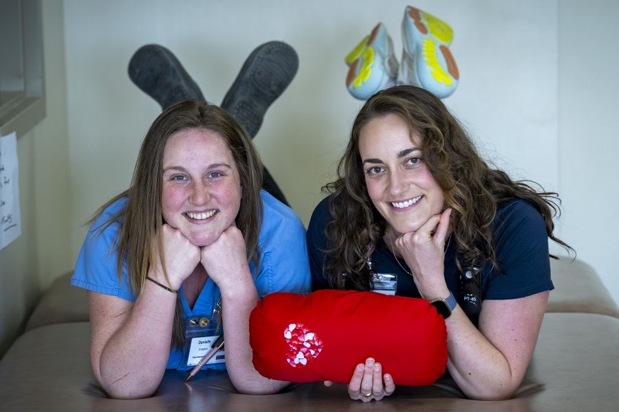 PeopleOfKHSC_Justine_White_10 Justine White and physiotherapy assistant Danielle Knapton are laying side-by-side with their feet crossed in the air, holding a cardiac surgery pillow. White has shoulder length, medium brown, wavy hair and brown eyes. She’s wearing a navy blue shirt with her KHSC ID badge. Knapton has straight medium brown hair and blue eyes. She’s wearing light blue scrubs with her KHSC ID badge.