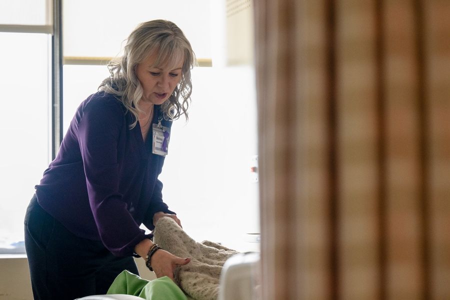Melissa Meisner is attending to a patient at the bedside. She’s standing at the end of the bed, holding a patient’s ankle. The patient is hidden behind a curtain. Meisner has blue eyes, shoulder length, silver hair and is wearing a bright purple blouse.