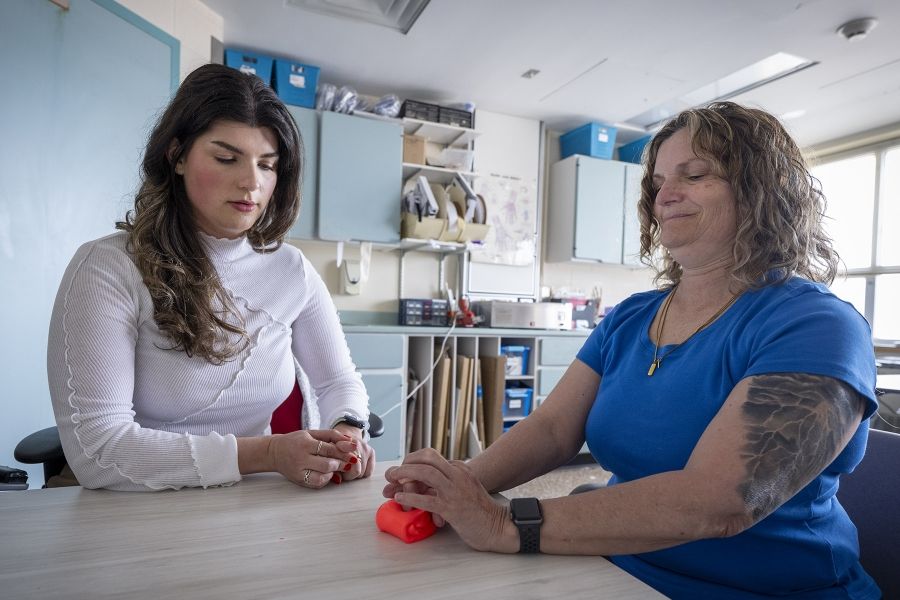 PeopleOfKHSC_Andreea_Bocicariu_3 Andreea Bocicariu is seated on a table, next to a patient in the Orthopedics and Plastics Clinic. The patient is doing a hand exercise with a bright orange tool. Bocicariu is observing her patient and looking directly at her hands.