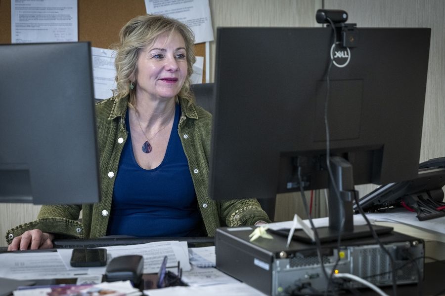 Christine Hennigar is working at her desk, looking at computer screen. She has light blonde, wavy hair, blue eyes and is wearing a navy blue top with an army green jacket.