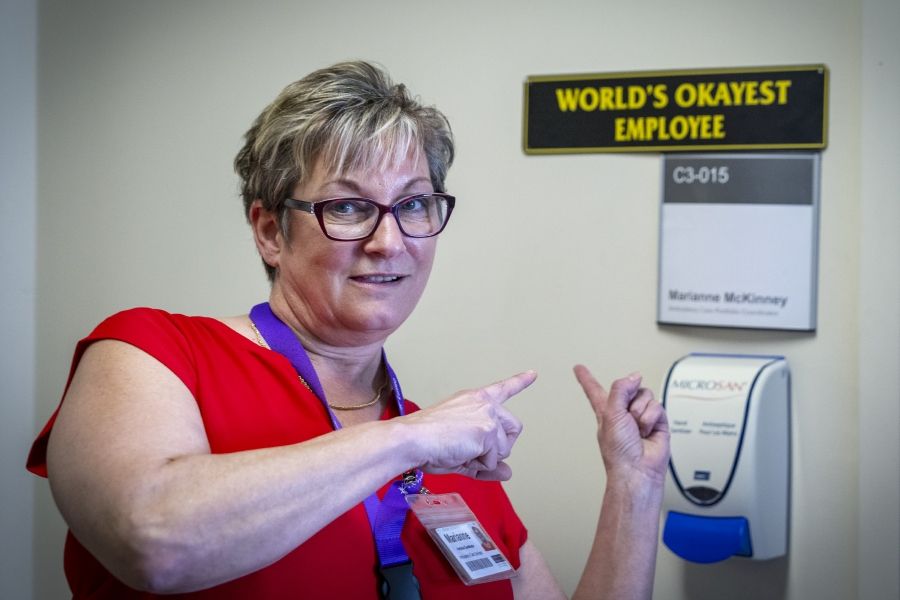 PeopleOfKHSC_Marianne_McKinney_6 A shot of Marianne McKinney standing outside her office pointing to a sign that says “World’s Okayest Employee.” McKinney has short dirty blonde hair, blue eyes, wears glasses and is dressed in a bright red blouse.