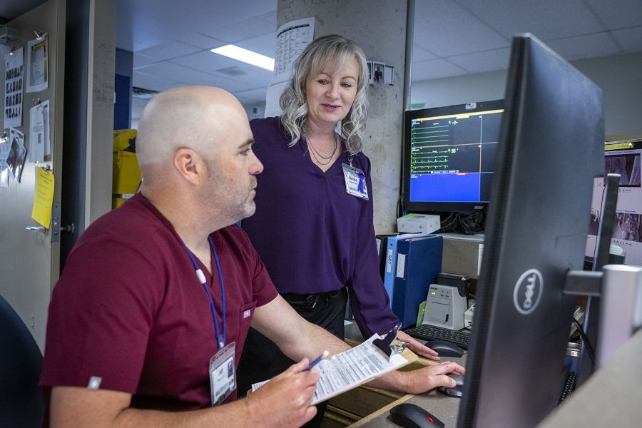 Melissa Meisner is standing behind a care desk, chatting with a colleague who is sitting down. They’re discussing what they’re seeing on nearby monitors. Meisner has blue eyes, shoulder length, silver hair and is wearing a bright purple blouse.