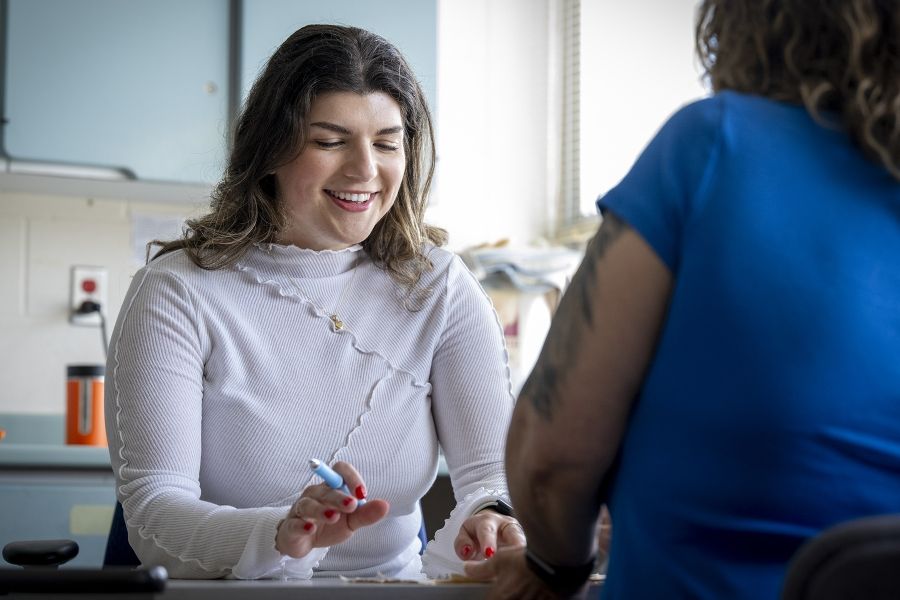 PeopleOfKHSC_Andreea_Bocicariu_5 Andreea Bocicariu is seated on a table across from her patient, whose back is to the camera, in the Orthopedics and Plastics Clinic. Bocicariu is smiling, while looking down at some paperwork with a pen in her hand.
