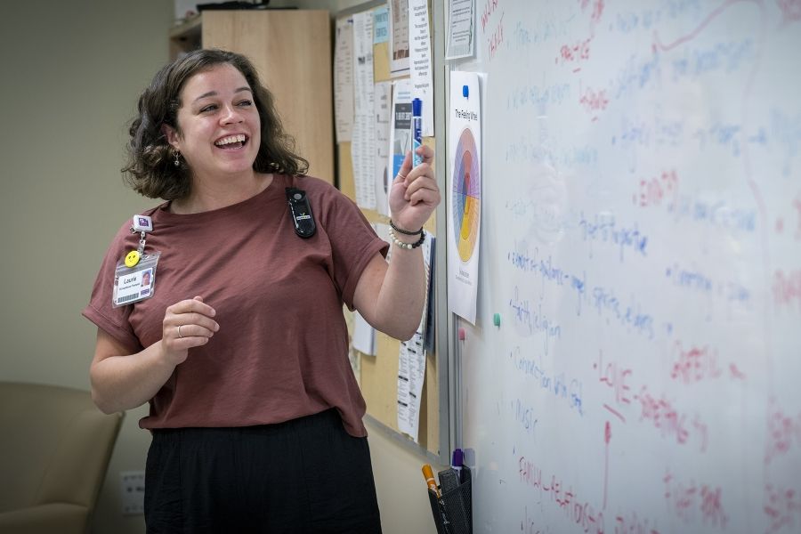 Laurie Hill is standing in front of a white board with a dry erase marker in her hand, and smiling. She has short brown wavy hair and is wearing a dusty rose blouse and black pants.