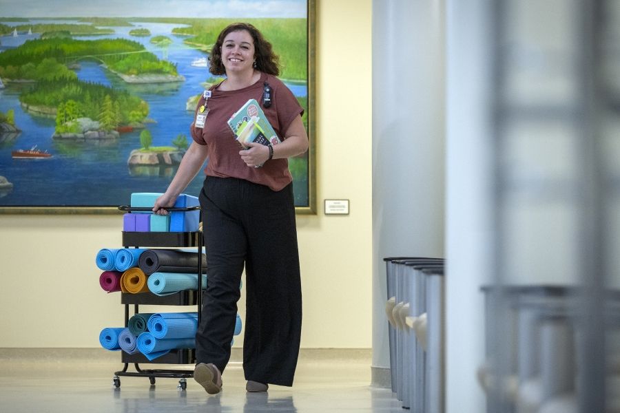 Laurie Hill is walking down a hallways, pulling a cart filled with yoga mats and blocks. She’s also holding a handful of notebooks in her arm. She has short brown wavy hair and is wearing a dusty rose blouse and black pants.