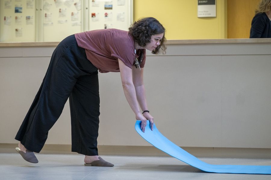 Laurie Hill is unrolling a blue yoga mat and placing it on the floor. She has short brown wavy hair and is wearing a dusty rose blouse and black pants.