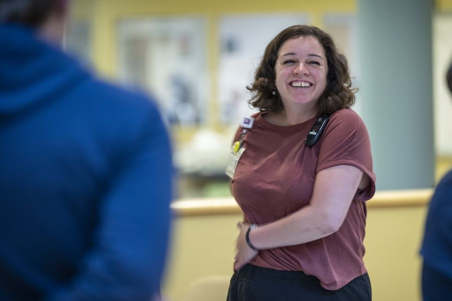 Laurie Hill is photographed leading her mindful movement group. Hill is standing and twisting her body to the side, with her arms placed on her hips. She has short brown wavy hair and is wearing a dusty rose blouse and black pants.