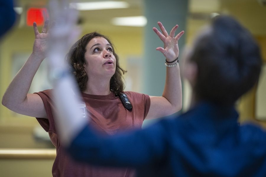Laurie Hill is photographed leading her mindful movement group. Hill is standing with her head slightly tilted back and her arms up in the air. She has short brown wavy hair and is wearing a dusty rose blouse and black pants.
