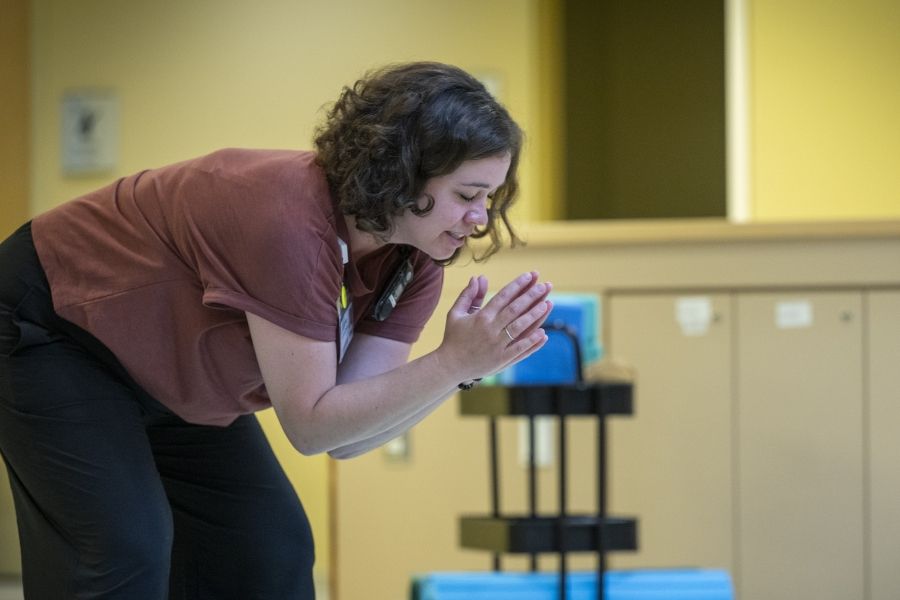 Laurie Hill is photographed leading her mindful movement group. Hill is bent over, with her arms and hands clasped together. She has short brown wavy hair and is wearing a dusty rose blouse and black pants.