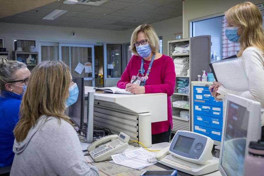 PeopleOfKHSC_Clare_Bowley_2 A wide shot of Clare Bowley who is standing at a care desk chatting with a few colleagues. Clare is taking notes in her black book. Clare has a blonde bob, blue eyes, has tortoise framed glasses on and a mask, and is wearing a pink sweater with her decorated KHSC lanyard on top.