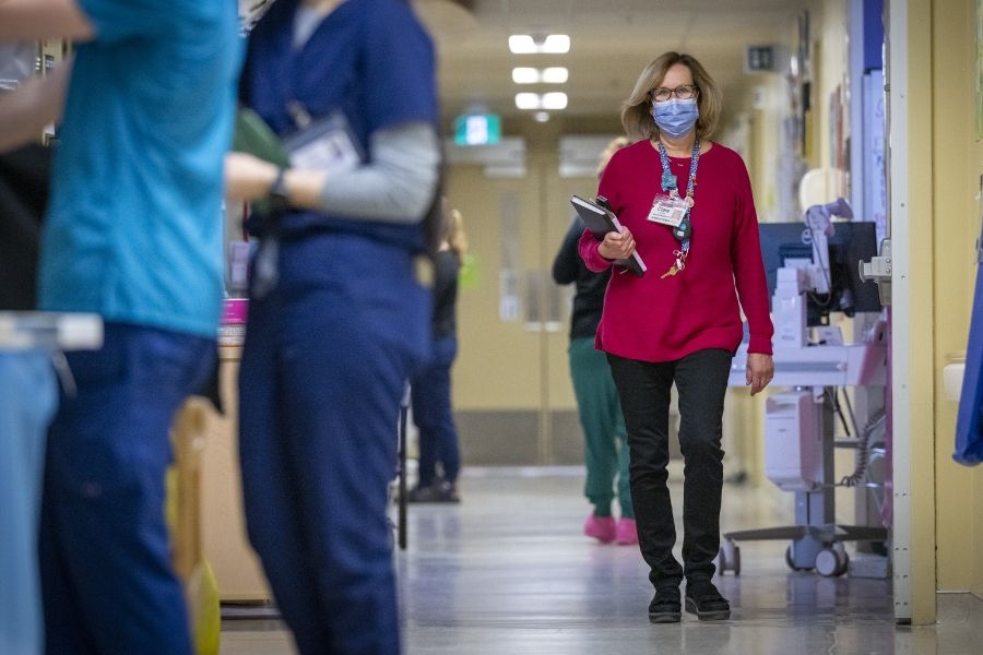PeopleOfKHSC_Clare_Bowley_3 A wide shot of Clare Bowley walking down a unit hallway wearing a mask. Clare is holding her black book and calendar. Clare has a blonde bob, blue eyes, has tortoise framed glasses on, and is wearing a pink sweater with her decorated KHSC lanyard on top.