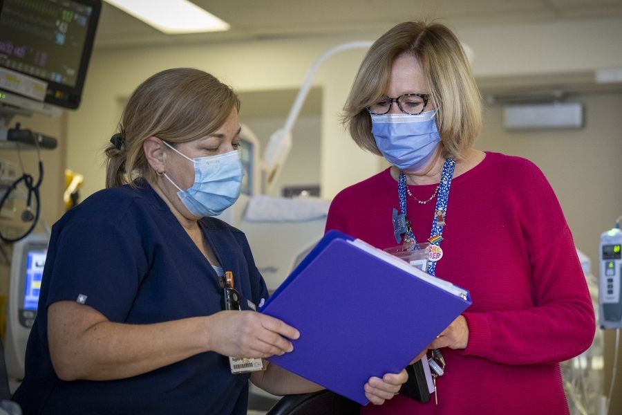 PeopleOfKHSC_Clare_Bowley_6 A wide shot of Clare Bowley going over a blue patient binder with the charge nurse in the Neonatal Intensive Care Unit. Clare has a blonde bob, blue eyes, has tortoise framed glasses on and a mask, and is wearing a pink sweater with her decorated KHSC lanyard on top. The charge nurse has blonde hair which is tied in a ponytail and is wearing a mask and navy blue scrubs.