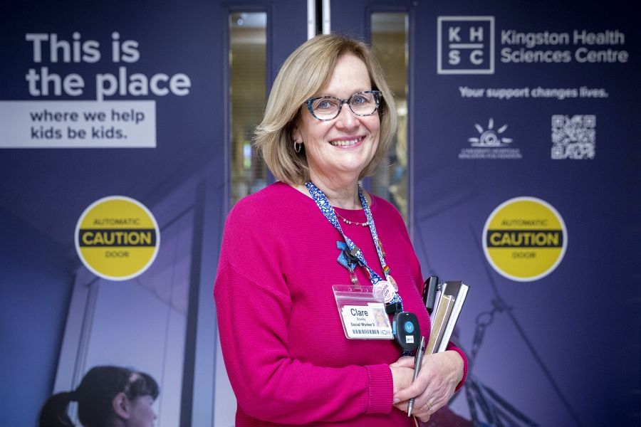 PeopleOfKHSC_Clare_Bowley_7 A wide shot of Clare Bowley standing outside the Pediatric unit and looking directly into the camera, smiling. Clare has a blonde bob, blue eyes, has tortoise framed glasses on, and is wearing a pink sweater with her decorated KHSC lanyard on top.