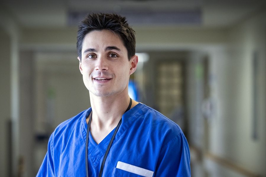 Evan Earl is pictured standing in a hallway at the Kingston General Hospital Site. He has short, dark, spiky hair, and brown eyes. He’s wearing bright blue scrubs.