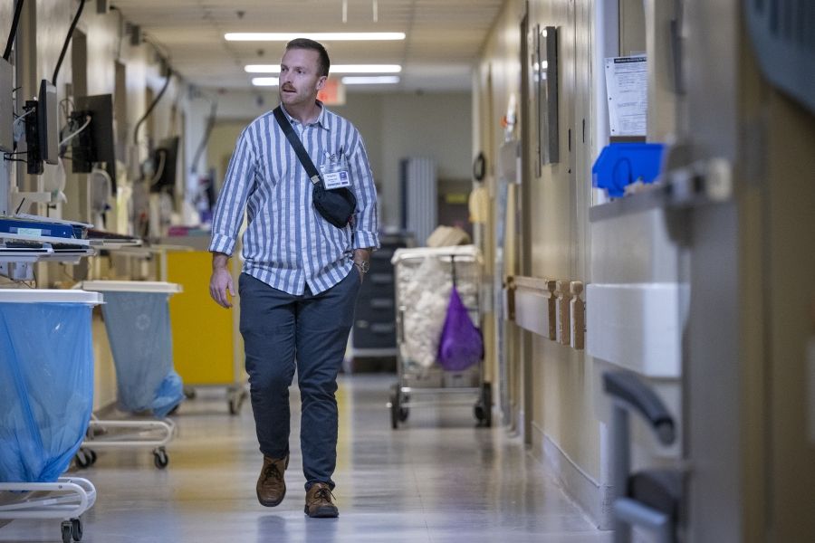 Khayman Wood is walking down a hallway at the Kingston General Hospital site. The hallway is lined with medical equipment, including carts with blue waste bags and monitors on the left. A yellow storage unit and other supplies are visible in the background.