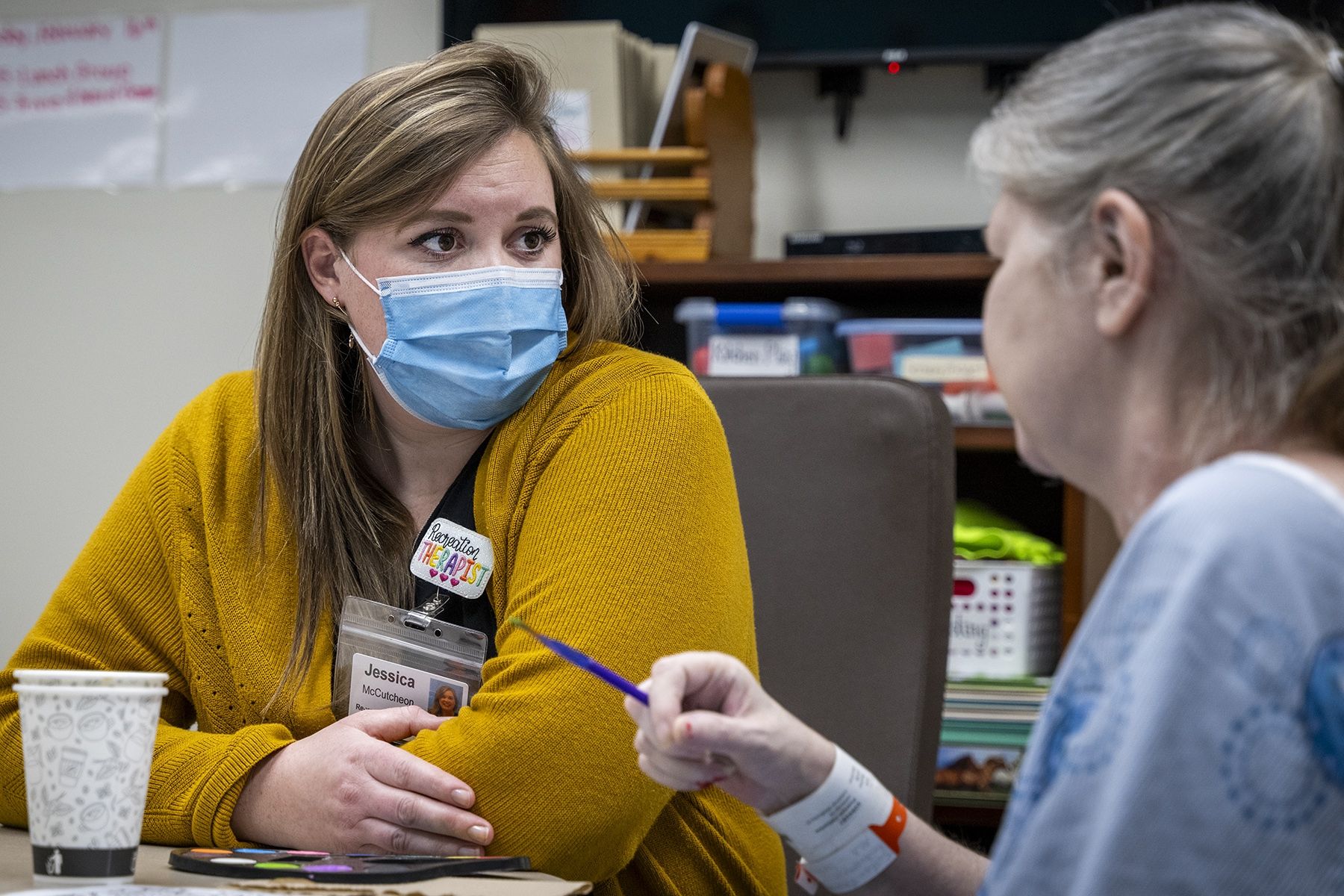 Jessica McCutcheon is seated next to an elderly female patient, watching her do arts and crafts. McCutcheon has dark blonde hair and brown eyes. She’s wearing a black top, mustard cardigan and a mask over her mouth and nose. 