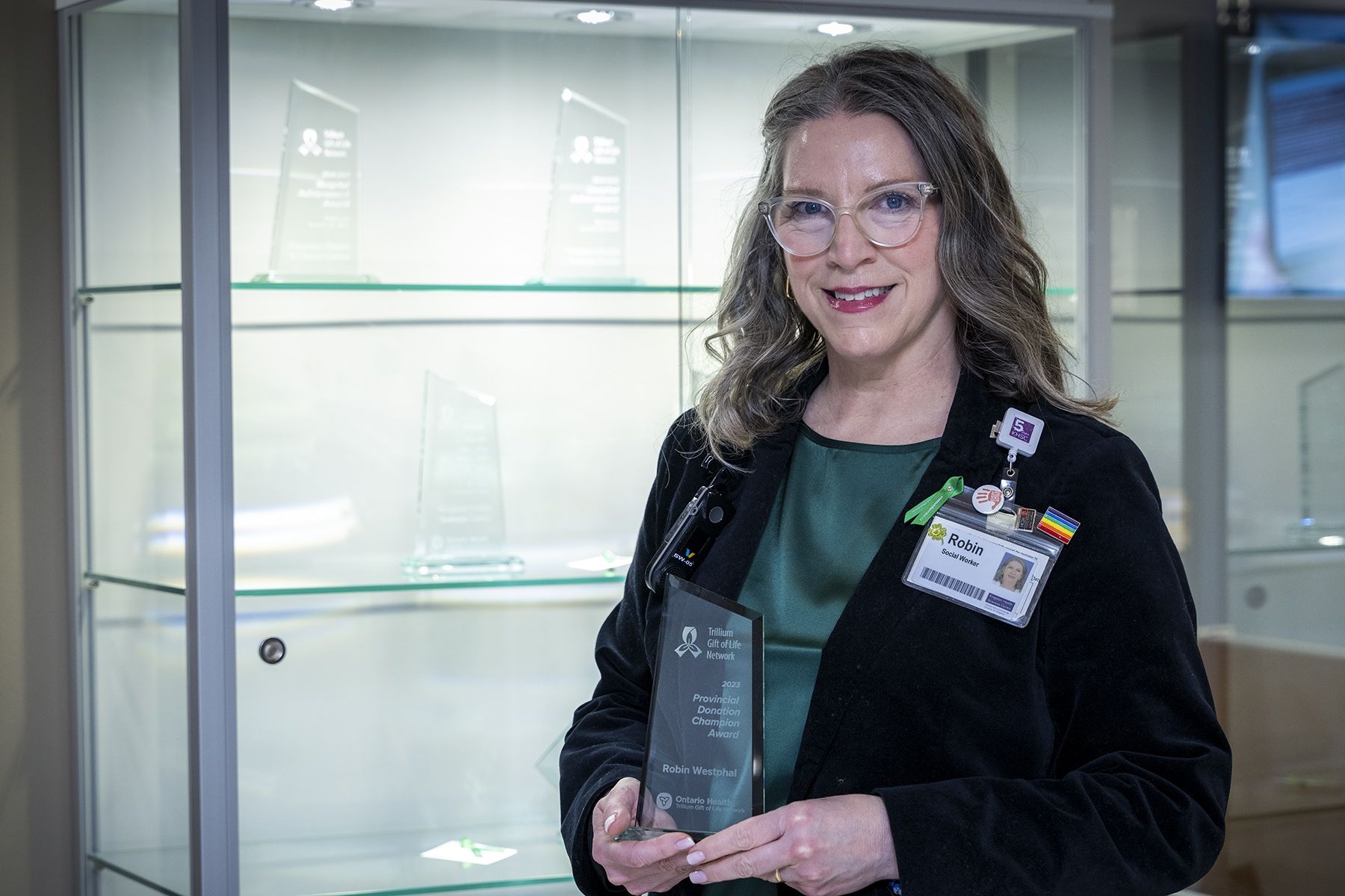 Robin Westphal is pictured standing in front of an awards display at Kingston General Hospital, holding her 2023 Provincial Donor Champion Award in her hands. Robin has dirty blonde hair with grey streaks, blue eyes and wears glasses. She’s wearing a green blouse with a black blazer on top.