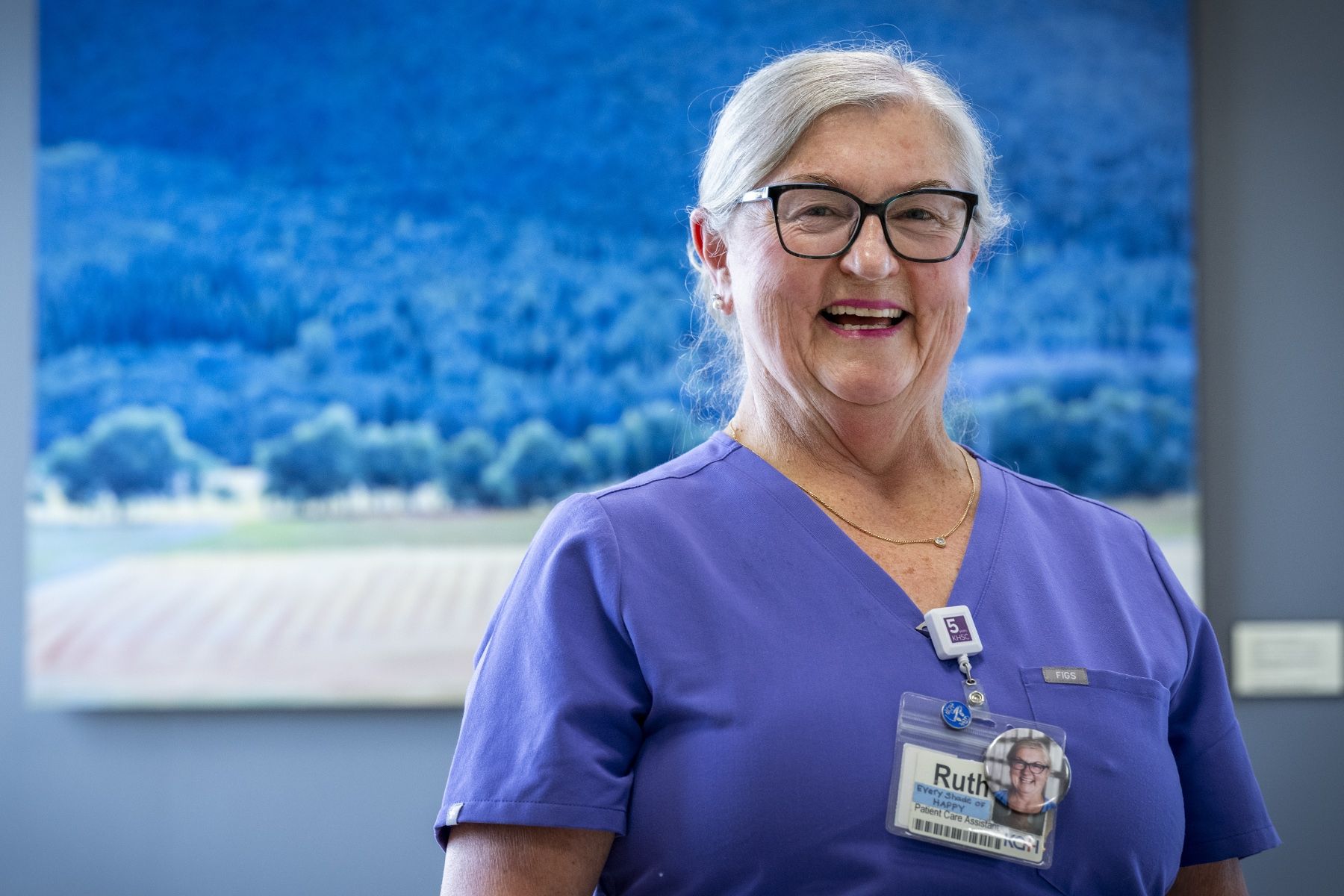 Ruth Bradshaw is pictured smiling directly at the camera while standing in a hallway at Kingston General Hospital. She has white/silver hair, pulled back into a bun, black framed glasses and is wearing blue scrubs.