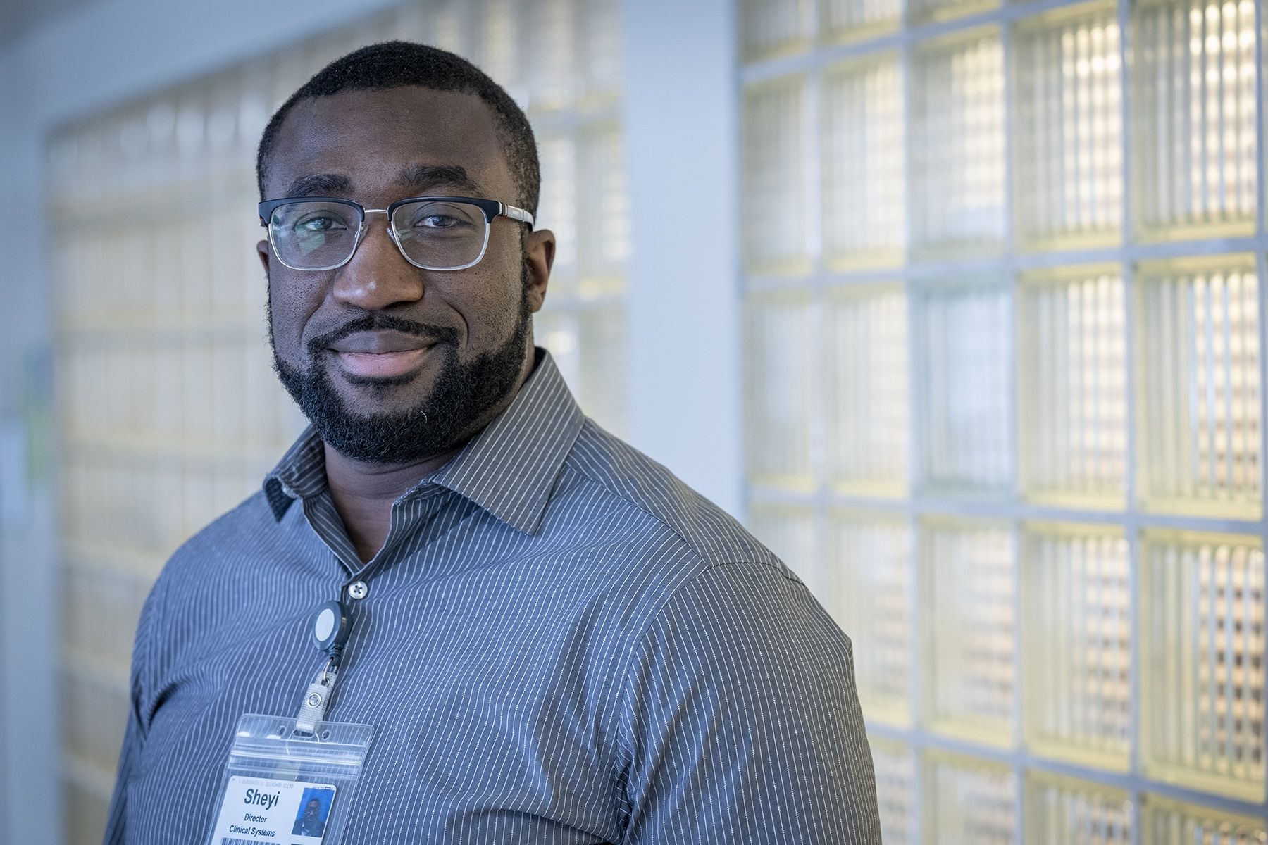 Sheyi Badmos is pictured standing in a hallway at the Kingston General Hospital Site. He’s Black, has short-dark hair, a beard, and wears glasses. He’s wearing a grey, pinstriped, button down shirt. 