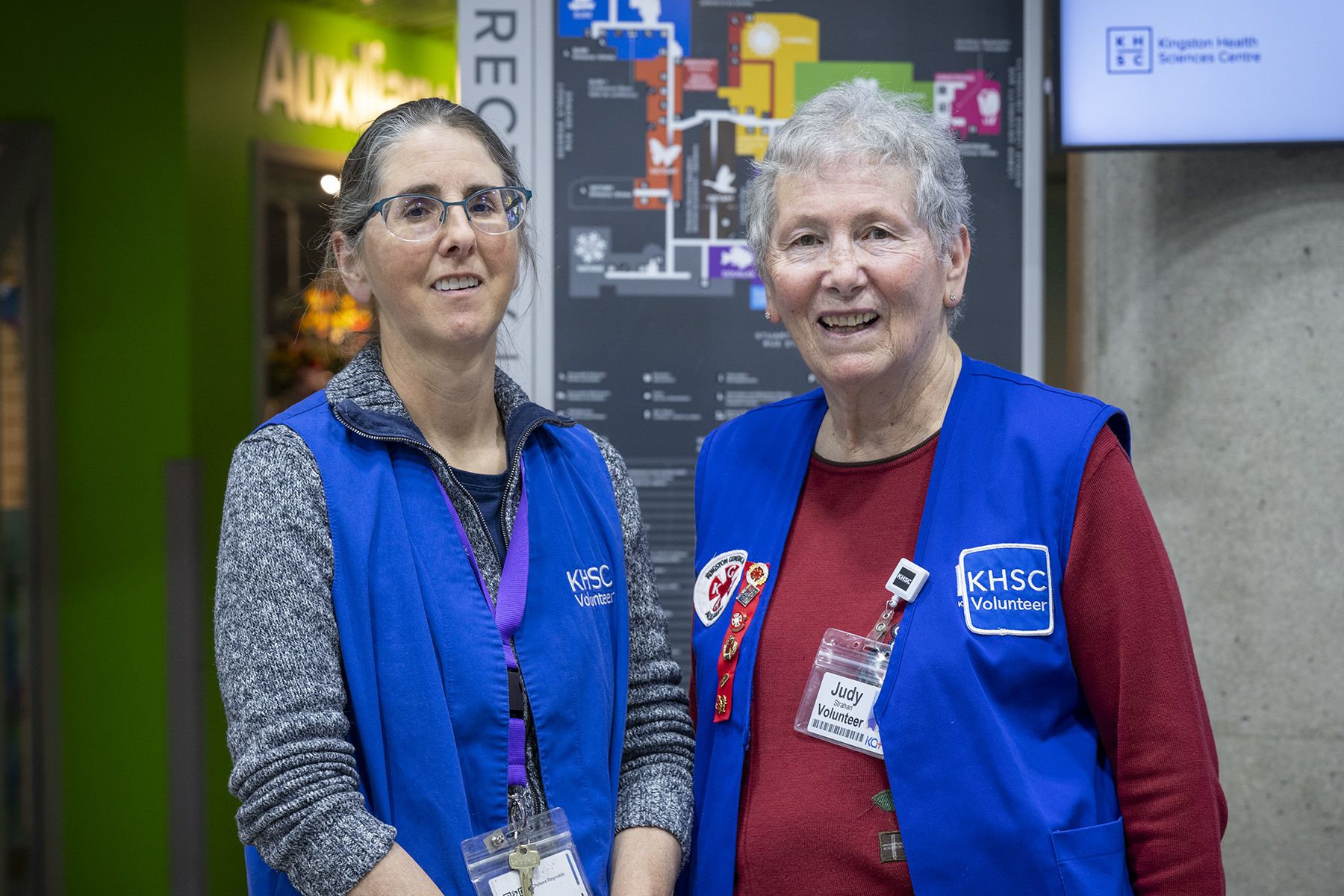 Two KHSC volunteers wearing blue vests stand side by side indoors in front of a hospital directory sign.