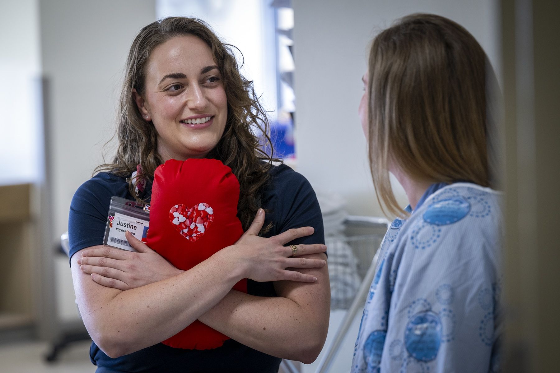 PeopleOfKHSC_Justine_White_3 Justine White is standing, hugging a cardiac surgery pillow. She’s smiling and looking directly at another person whose back is to the camera. She has shoulder length, medium brown, wavy hair and brown eyes. She’s wearing a navy blue shirt with her KHSC ID badge.