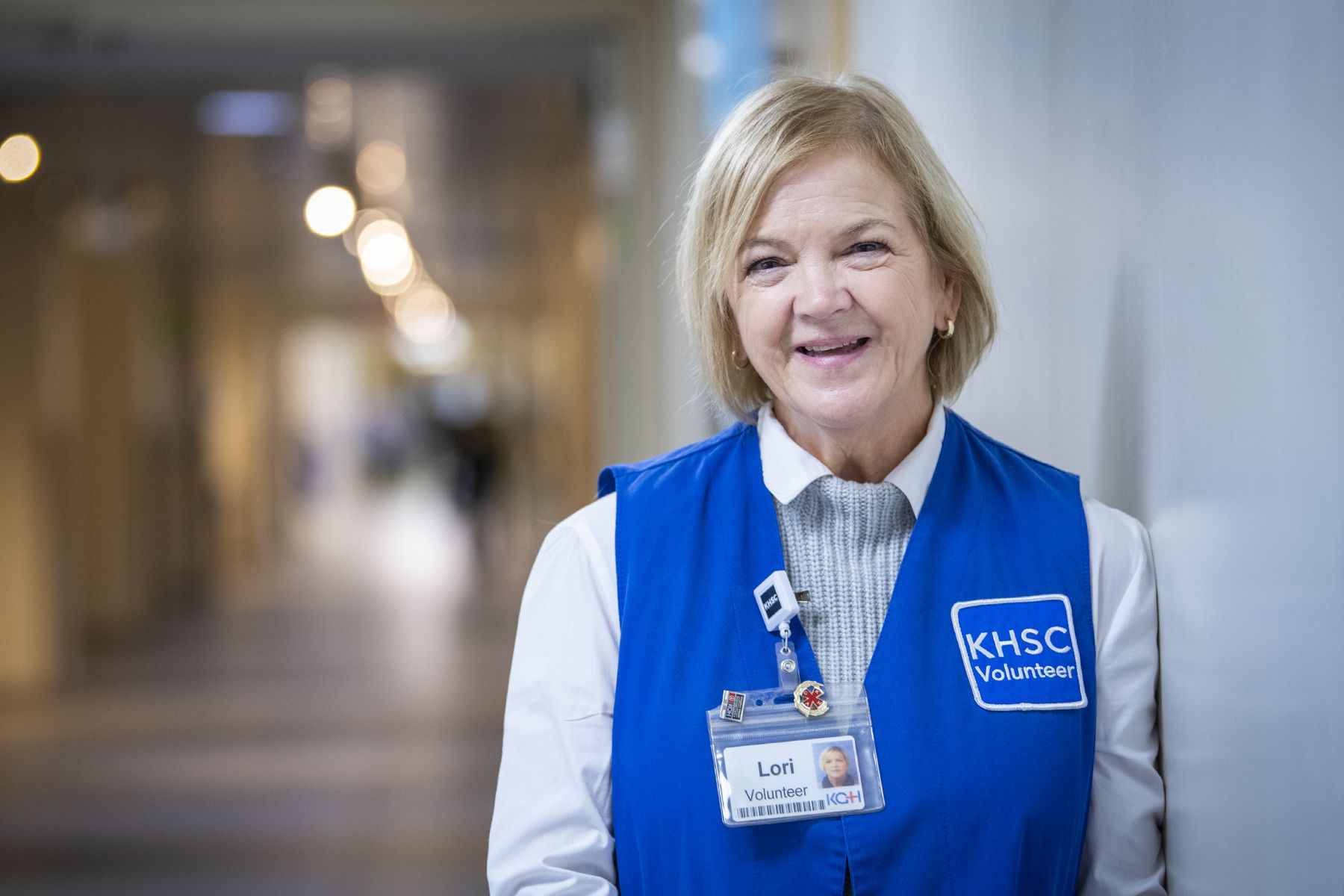 Hospital volunteer wearing a blue Kingston Health Sciences Centre volunteer vest and name badge, standing in a hospital hallway.