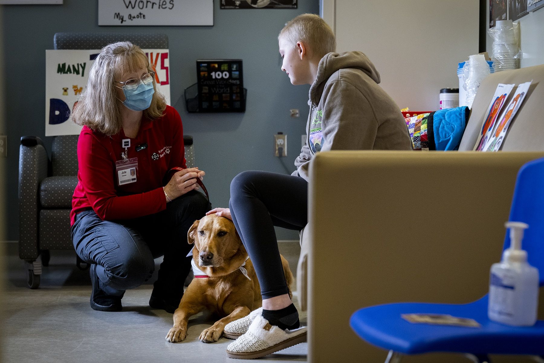 Julie Halle and Doc are sitting with a patient. Julie is sitting opposite the patient and chatting with her, while she pets Doc on the head as he lays on the floor.