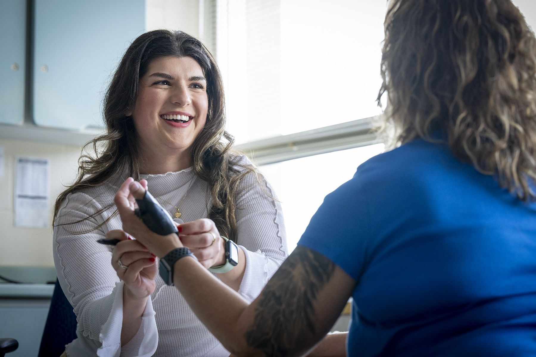 PeopleOfKHSC_Andreea_Bocicariu_8 Andreea Bocicariu is sitting at a table in the Orthopedics and Plastics Clinic, across from a patient whose back is to the camera. She’s smiling from ear to ear at her patient, while wrapping a splint on her hand.