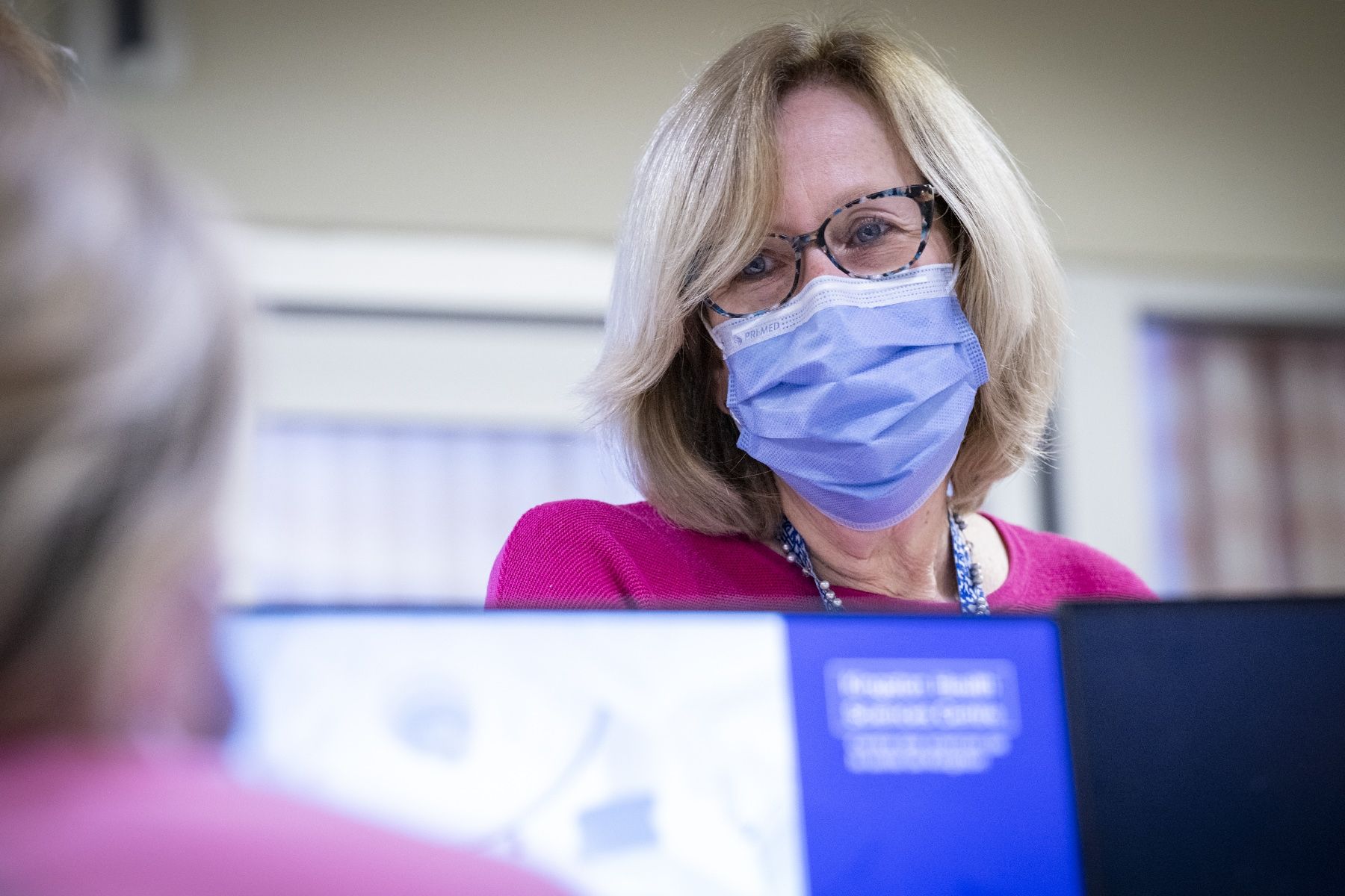 A close up shot of Clare Bowley who is standing at a care desk chatting with a colleague who is seated. Clare has a blonde bob, blue eyes, has tortoise framed glasses on and a mask, and is wearing a pink sweater.