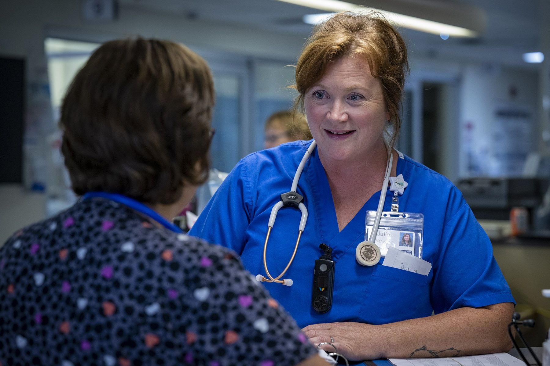 Julie Vanyolai is pictured at a care desk talking to a colleague whose back is turned to the camera. She has strawberry blonde/red hair, which is tied up in a bun, and blue eyes. She’s wearing blue scrubs and has a stethoscope around her neck. 