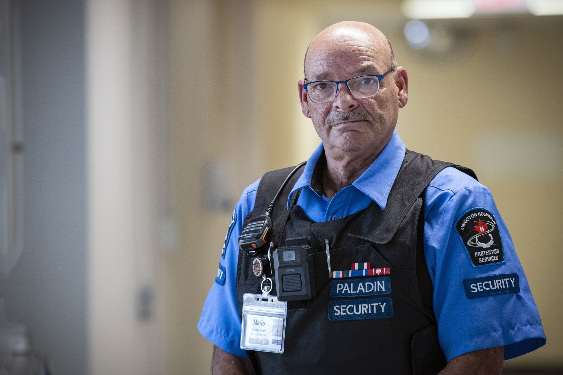Mario Lelievre is pictured standing in a hallway at Hotel Dieu Hospital. He has brown eyes, wears blue glasses and is bald. He’s wearing his Paladin Security uniform, which includes a blue collared, button down shirt, protective vest, radio, and ID badge.