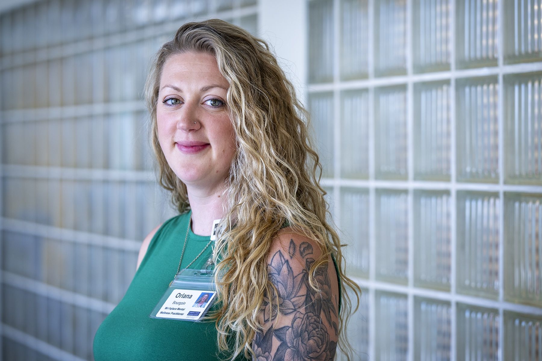 Orlana Bourgoin is standing in a hallway at the Kingston General Hospital site, looking directly in the camera. She has long, wavy, blonde hair, blue eyes and is wearing a bright green tank top. Her upper left arm is covered in flower tattoos.