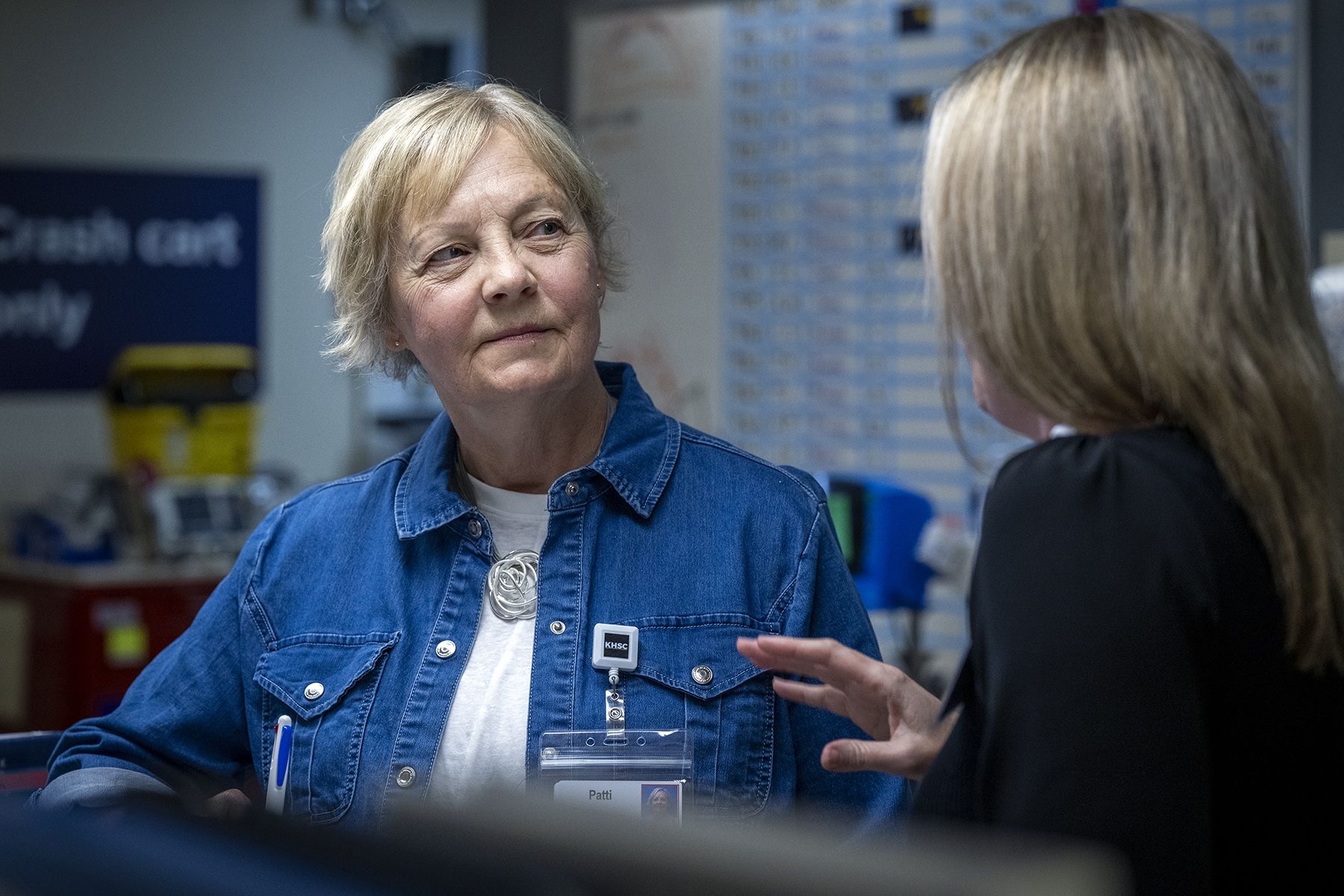 Patti Cox is pictured leaning against a care desk on a patient care unit, listening as a member of the care team speaks to her. Cox has blue eyes and light blonde hair styled in a pixie cut. She’s wearing a blue denim blouse on top of a white t-shirt. The staff member has their back to the camera, while Cox is facing the camera.