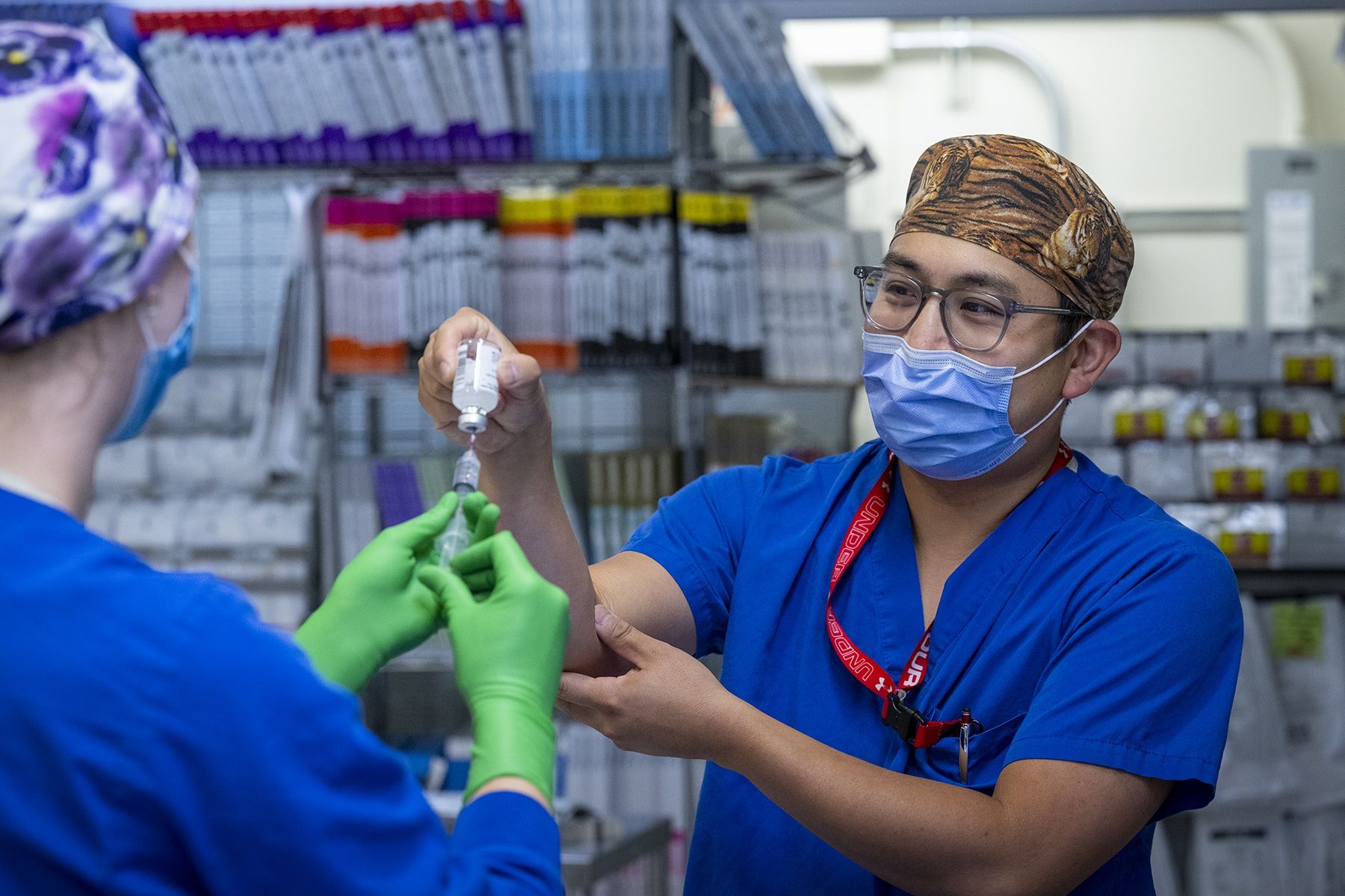 Warren Lee is pictured standing in a room filled with medical equipment. Lee is holding a vial while his colleague is using a syringe and needle to get the substance out of the vial. Lee is wearing bright, blue scrubs, tiger print scrub cap, a mask over his nose and mouth, and has grey, translucent glasses.