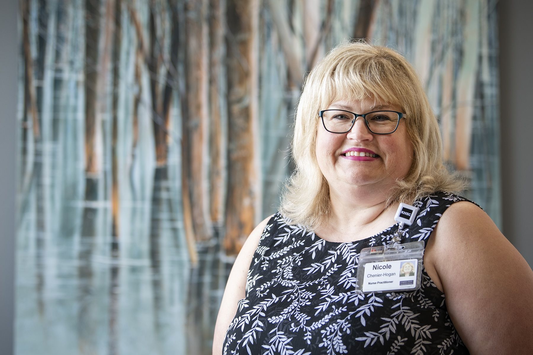 Nicole Chenier-Hogan is pictured standing in a hallway at Kingston General Hospital. She has blonde, shoulder-length hair, has glasses and is wearing a black blouse with white leaves. 