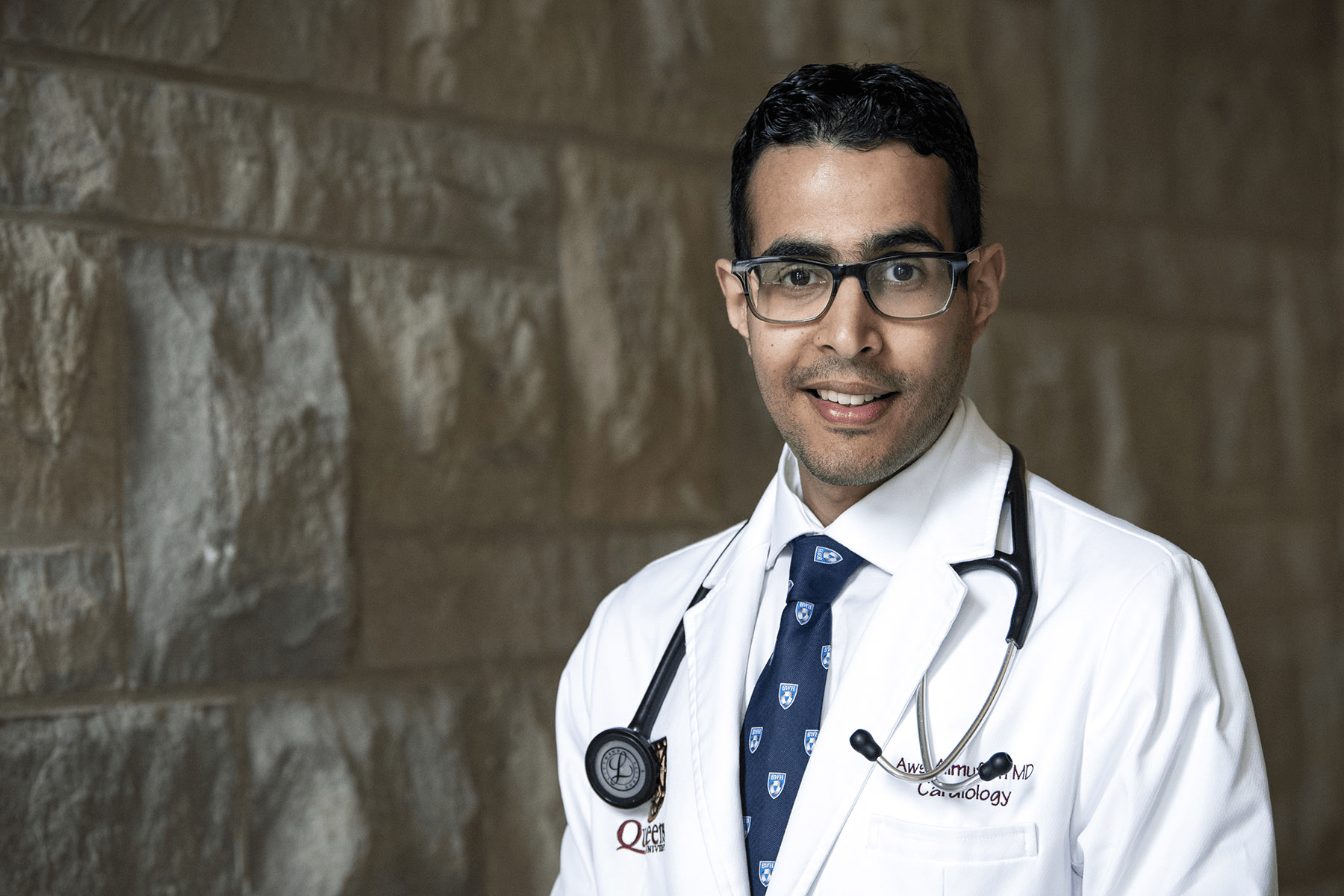 Dr. Aws Almufleh is pictured standing in a hallway at the Hotel Dieu Hospital site. He has short, dark hair, dark brown eyes, has glasses, and is pictured wearing a white laboratory coat. 